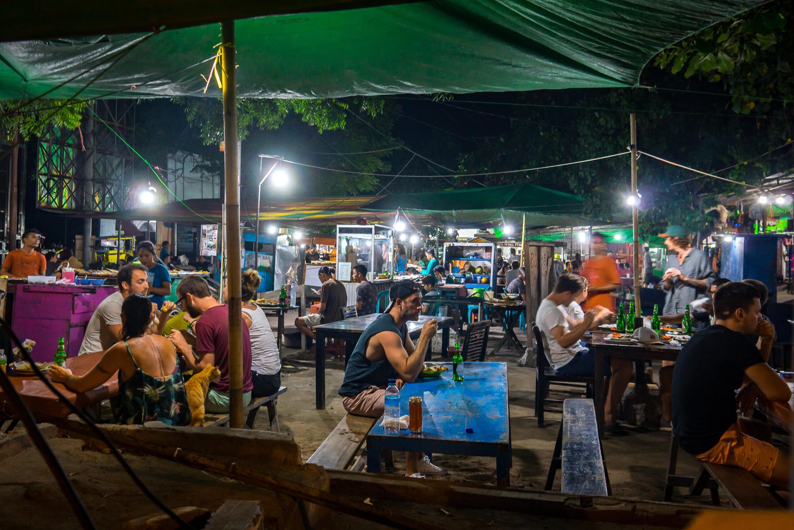 European turists and local residents sitting in the light of electrical bulbs at long tables having various streetfood in an indonesian nightmarket, a